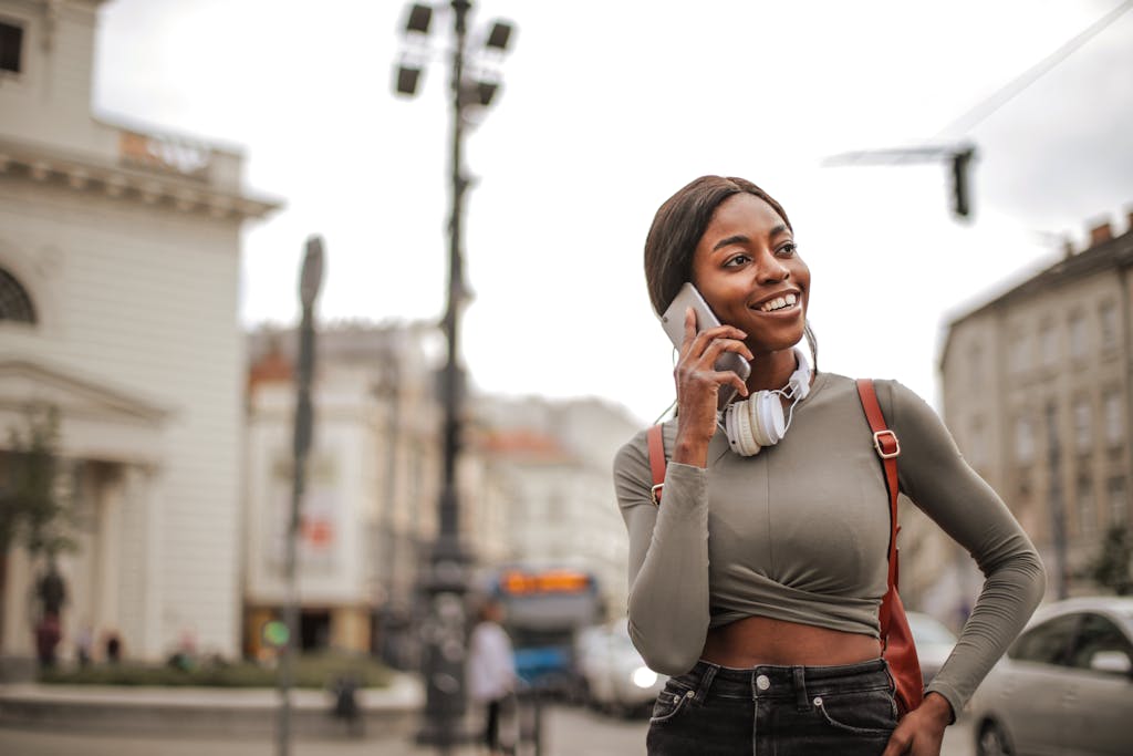 Selective Focus Photo of Smiling Woman in Gray Long Sleeve Shirt and Black Denim Jeans Standing While Talking on the Phone